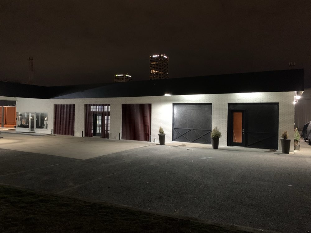 The 801 at 801 S Chester Street at night: a white brick building with a glass storefront, red slatted screens and dark barn-style doors, the downtown skyline behind it.
